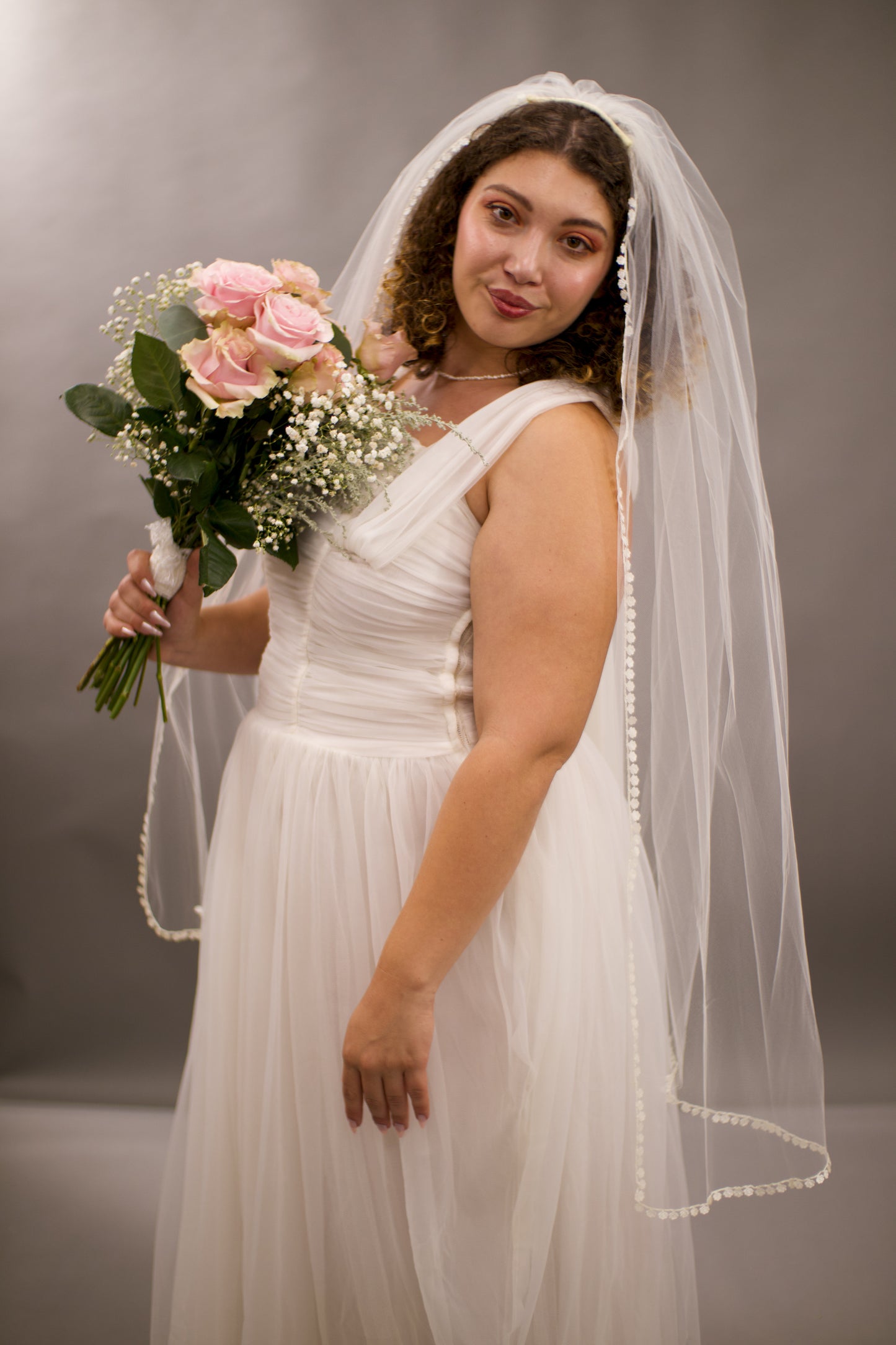 Woman in a white wedding dress with a veil holding a bouquet against a plain background